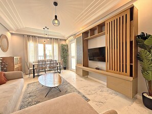 Uma sala de estar com sofá, mesa de jantar com cadeiras, televisão em um suporte de madeira, uma planta grande em vaso e um lustre.