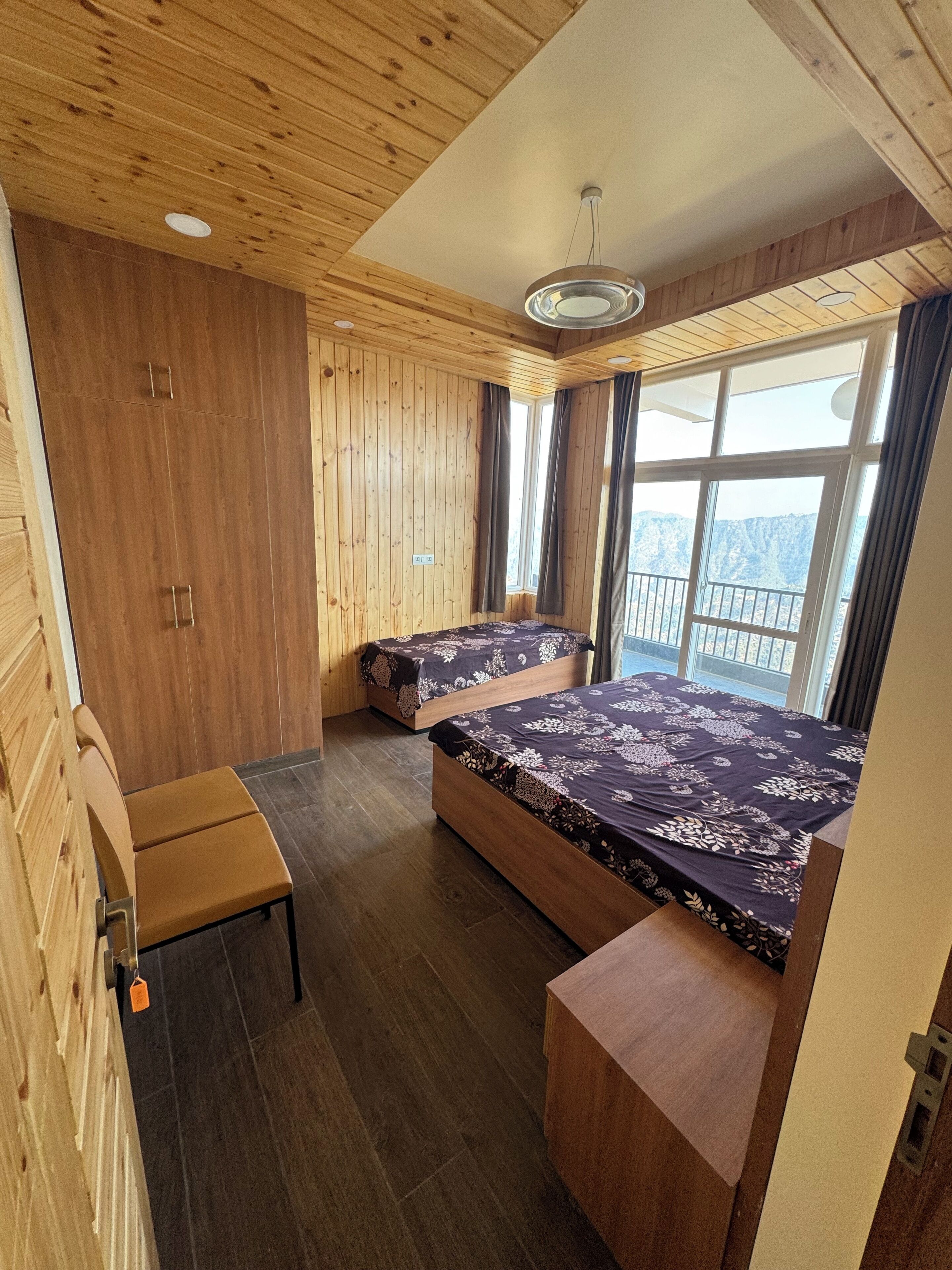 Quarto com paredes e teto de madeira, duas camas com colchas florais, um banco e uma janela com vista.