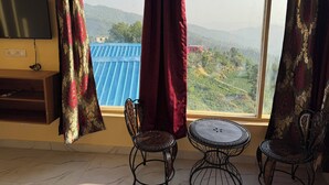 Una stanza con TV, due poltrone, un tavolino e un'ampia finestra che si affaccia su un edificio con tetto blu e delle colline.
