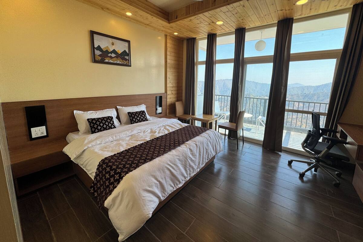 A hotel room with a large bed, a desk, a chair, and a balcony with a view of the mountains.