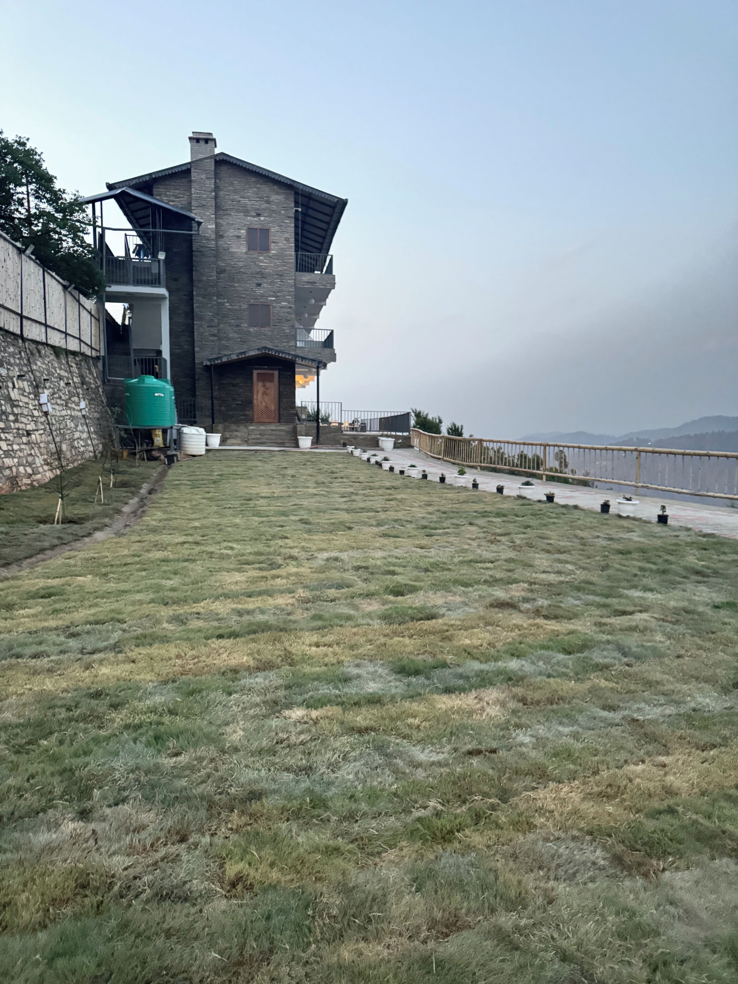 Un edificio moderno con fachada de piedra, un tanque de agua verde y una zona ajardinada con plantas en macetas.