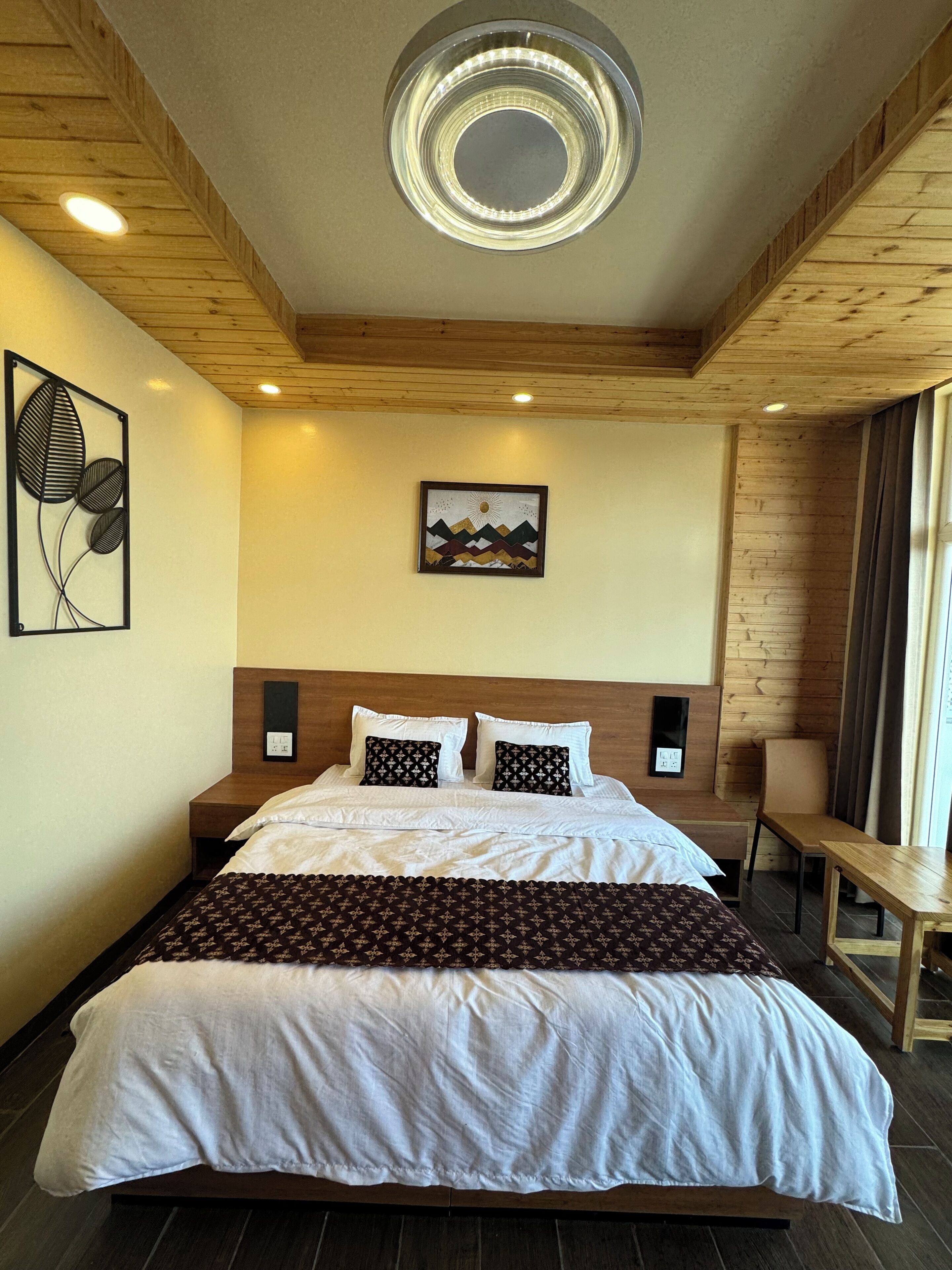 A bedroom with a wooden ceiling, a bed with white and patterned bedding, a wall-mounted artwork, and a bedside table.