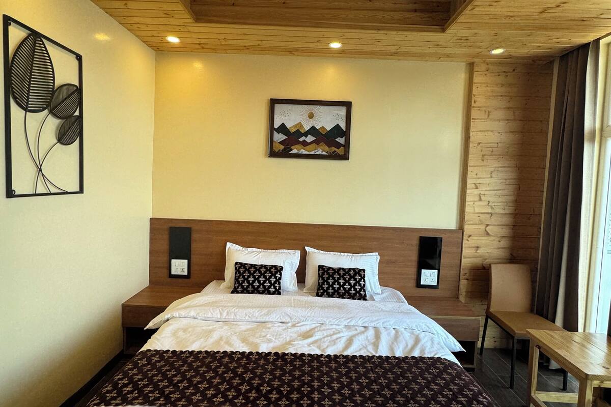 A bedroom with a wooden ceiling, a bed with white and patterned bedding, a wall-mounted artwork, and a bedside table.