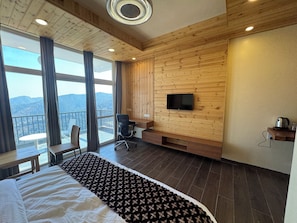 A modern hotel room with a large bed, a TV mounted on the wooden wall, a balcony with a view of the mountains, and a small desk area. - Ivaan Belle-Vue (Dhari)