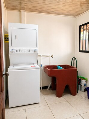 Een kleine wasruimte met een voorlader wasmachine en droger, een klein wasbakje met een slang, en een rode emmer.