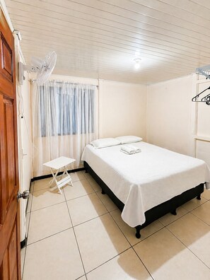 A bedroom with a bed, a small table, a fan, and a window with curtains. - Starlink Jungle Retreat | Eco Living + Fast WiFi (Cahuita)