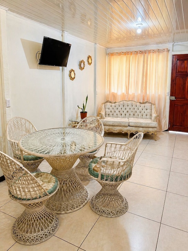 A living room with a round glass table, wicker chairs, a sofa, a TV mounted on the wall, and a potted plant.
