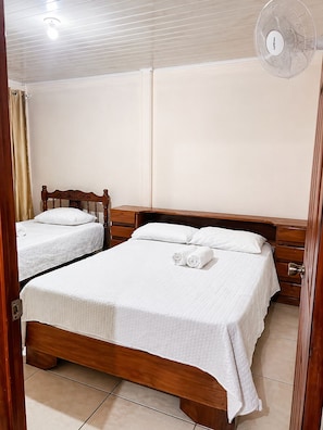 A hotel room with two beds, a wooden headboard, a bedside table, a wall-mounted fan, and a door with a curtain.