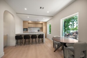 Una cocina moderna con una isla central, taburetes de bar, una mesa de comedor y un ventanal con vistas a la vegetación.