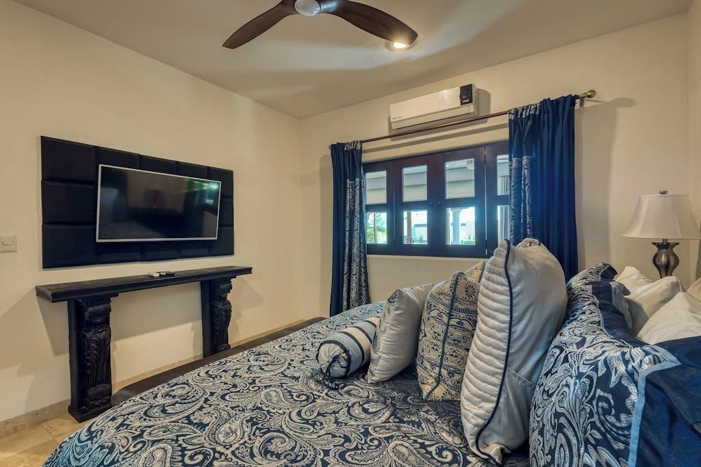 A bedroom with a bed, a TV mounted on the wall, a window with curtains, and a ceiling fan.