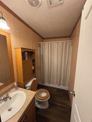 Un baño pequeño con inodoro de madera, lavamanos blanco, espejo, cortina de ducha y rejilla de ventilación en el techo.
