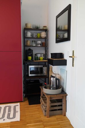 Ett kompakt kök med en mikrovågsugn, brödrost och kaffebryggare på hyllor, ett litet träbord med en vattenkokare och en röd dörr.