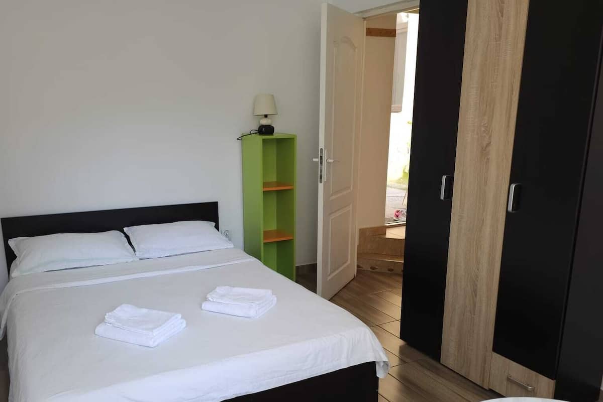 Un lit bien fait, avec du linge de lit blanc, une étagère verte, une armoire noire et une table ronde recouverte d’une nappe blanche.