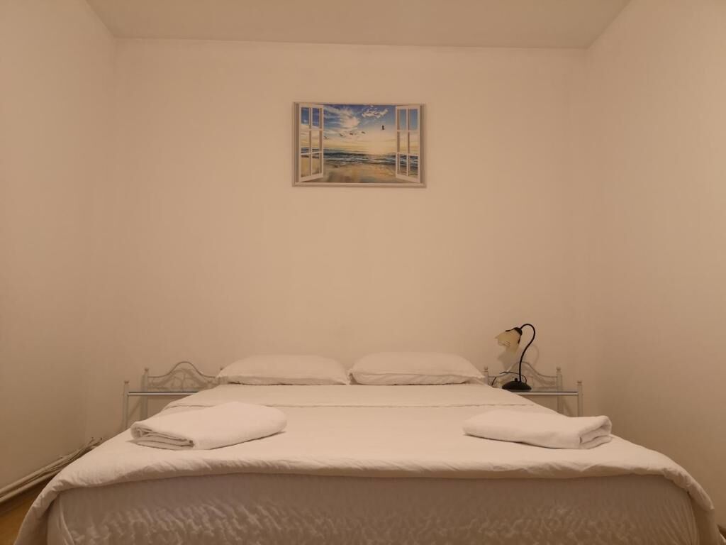 A bedroom with a bed, white linens, a nightstand, a lamp, and a painting of a beach scene.