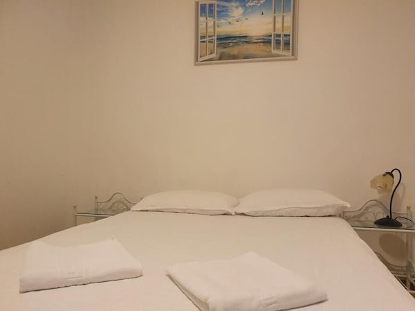 Una cama con sábanas blancas, una mesita de noche con una lámpara y un cuadro de una playa.