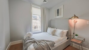 A neatly arranged bedroom with a bed, bedside table, lamp, and a window with a view of buildings. - Spacious 1bed/1bath in Back Bay (Boston)