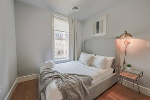 Un dormitorio ordenado con cama, mesita de noche, lámpara y ventana con vistas a edificios.