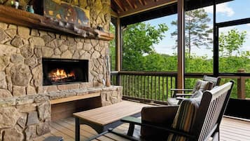 A stone fireplace with a fire, wooden deck seating, and a view of trees.
