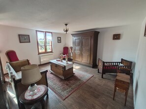 Una habitación con piso de madera, una silla roja, un armario de madera, una mesita con una lámpara y una cuna.