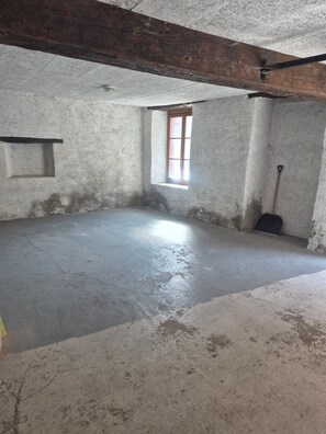 Una habitación con piso de concreto, una ventana con contraventanas de madera, una escoba apoyada en la pared y un estante pequeño.