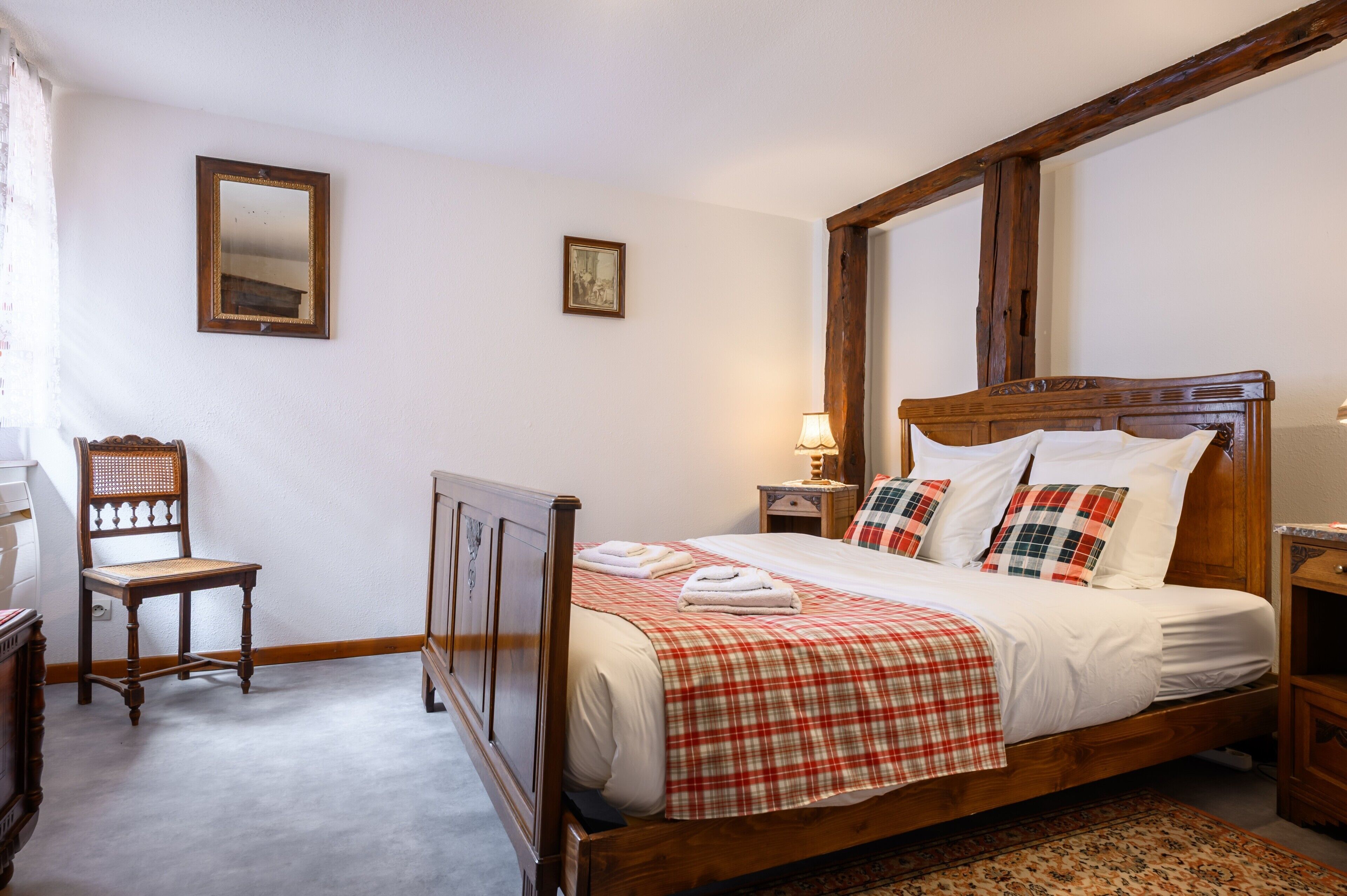A wooden bed with a plaid bedspread, a wooden nightstand with a lamp, a wooden chair, and a small table with a lamp.