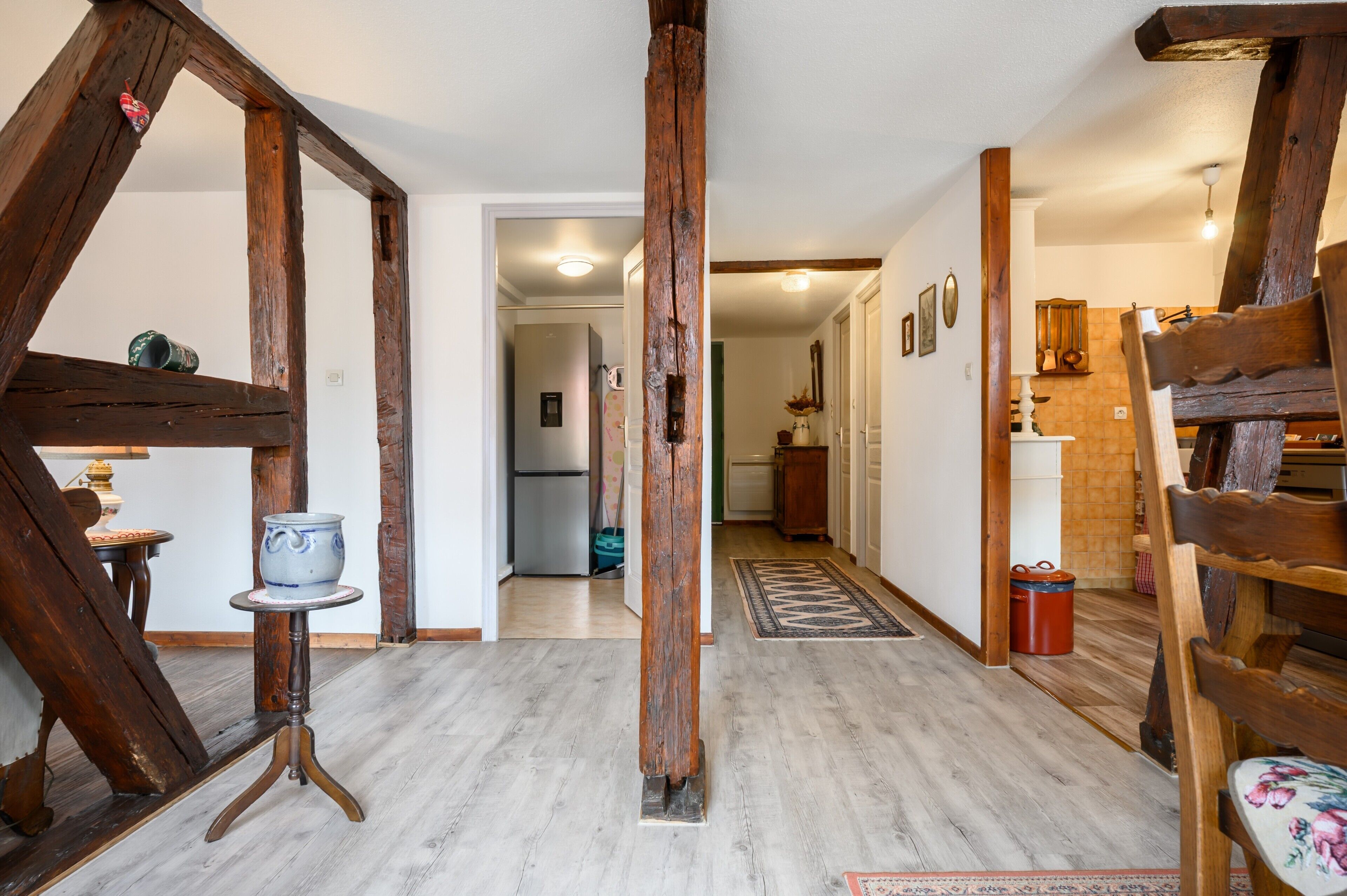 A rustic interior with exposed wooden beams, a wooden bench, a small table with a vase, and a rug leading to a kitchen area.