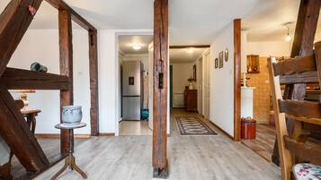 A rustic interior with exposed wooden beams, a wooden bench, a small table with a vase, and a rug leading to a kitchen area.