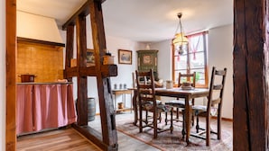 Een landelijke eethoek met een houten tafel en stoelen, een hanglamp en uitzicht door een raam met gordijnen.