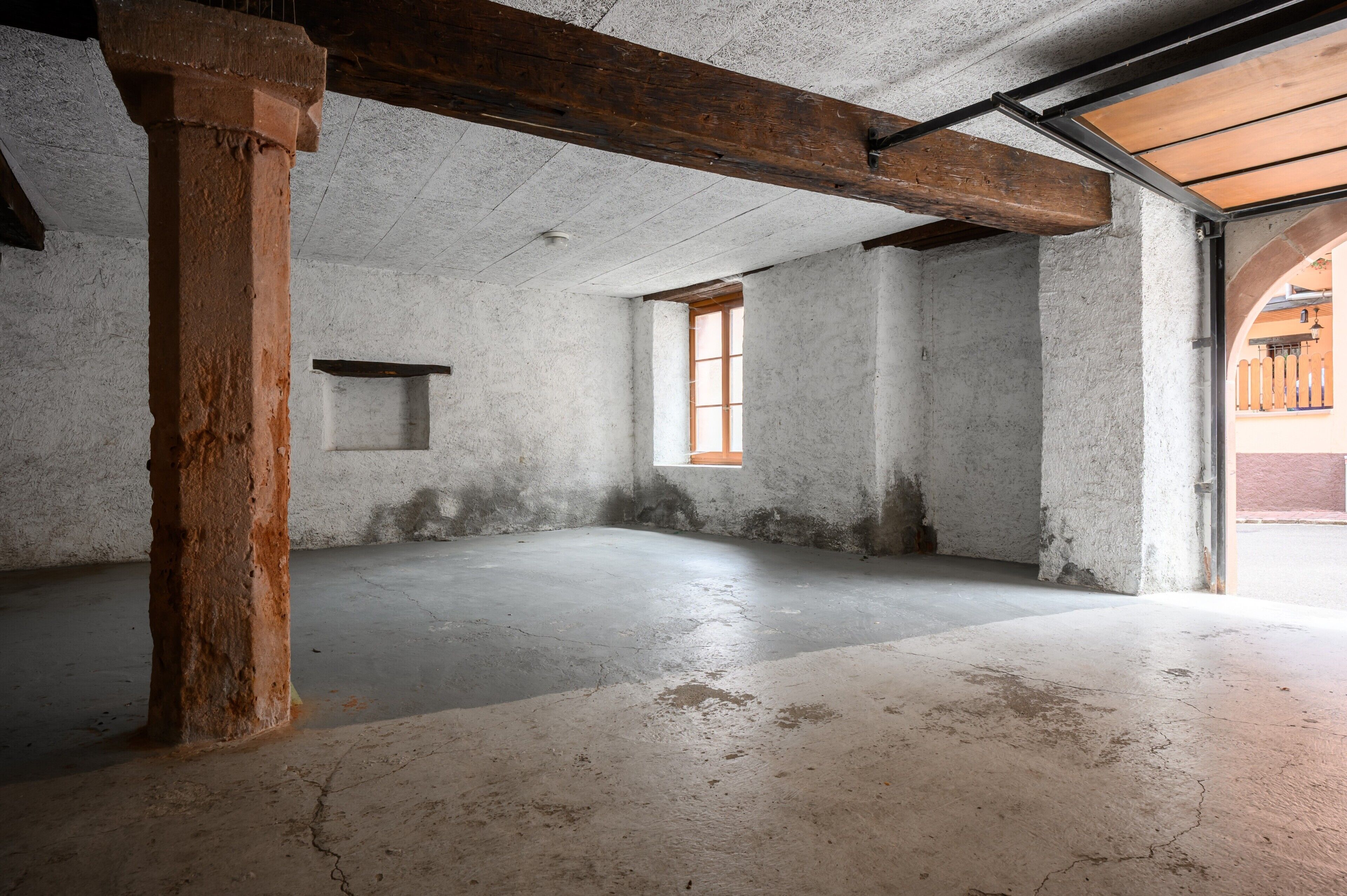 Un interior con paredes de concreto a la vista, techo con vigas de madera y una única ventana.