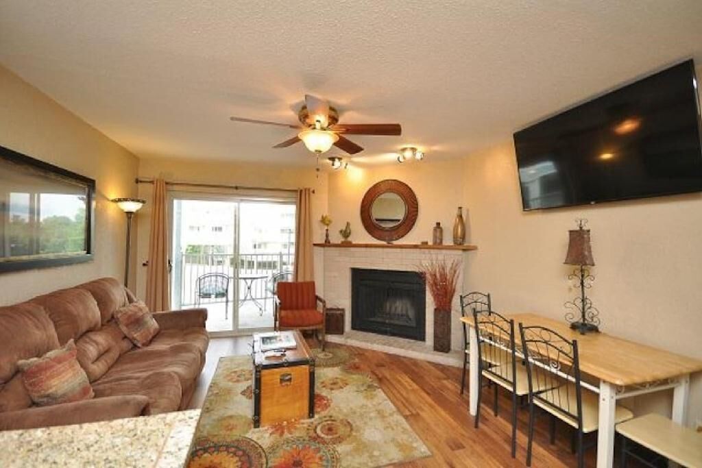 A living room with a sofa, a coffee table, a fireplace, a dining table with chairs, a ceiling fan, and a TV mounted on the wall.