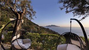 Two wicker swing chairs with cushions, a stone pathway, a tree, and a view of the sea. - Luxury Villa with Private Pool, Capri Views and Panoramic Rooftop (Campania)