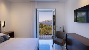 A hotel room with a bed, a chair, a desk, a TV, and a balcony with a view of the sea and mountains. - Luxury Villa with Private Pool, Capri Views and Panoramic Rooftop (Campania)
