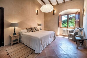 Un dormitorio con cama, lámparas de noche, una ventana con vistas, una silla de mimbre y una mesa con un florero.