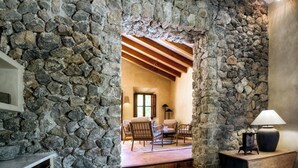 Un intérieur aux murs de pierre, avec une cheminée, un escalier menant à un espace détente doté de fauteuils, et une lampe posée sur une table en bois.
