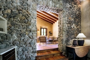 A stone-walled interior with a fireplace, a staircase leading to a seating area with chairs, and a lamp on a wooden table.