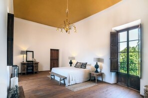A bedroom with a large bed, two bedside tables, a chandelier, a wooden bench, a wooden door, and a window with a view of greenery.