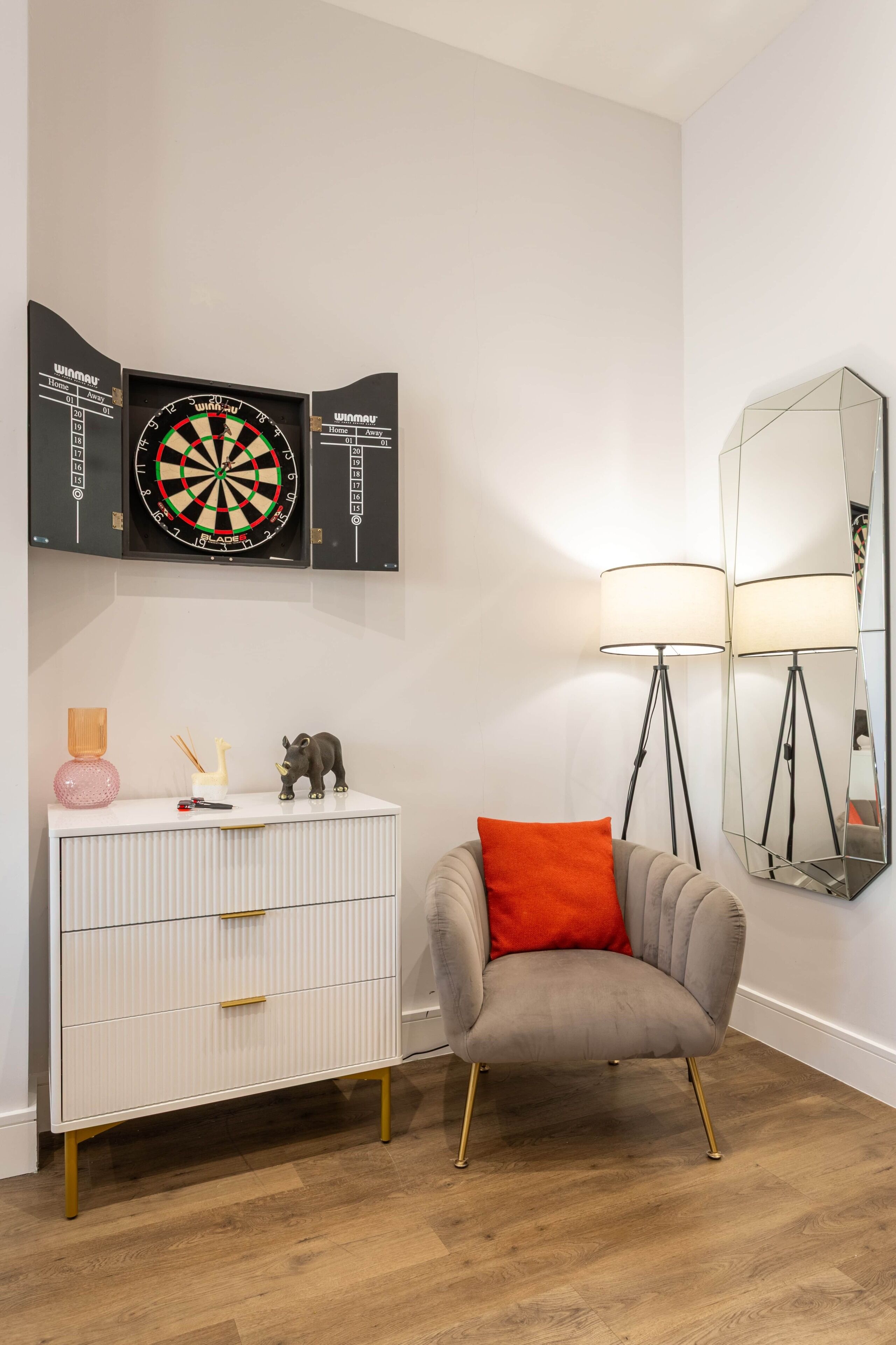 Ein Zimmer mit Dartscheibe, einer Lampenschirm aus Spiegelglas, einer weißen Kommode und einem grauen Sessel mit orangefarbenem Kissen.