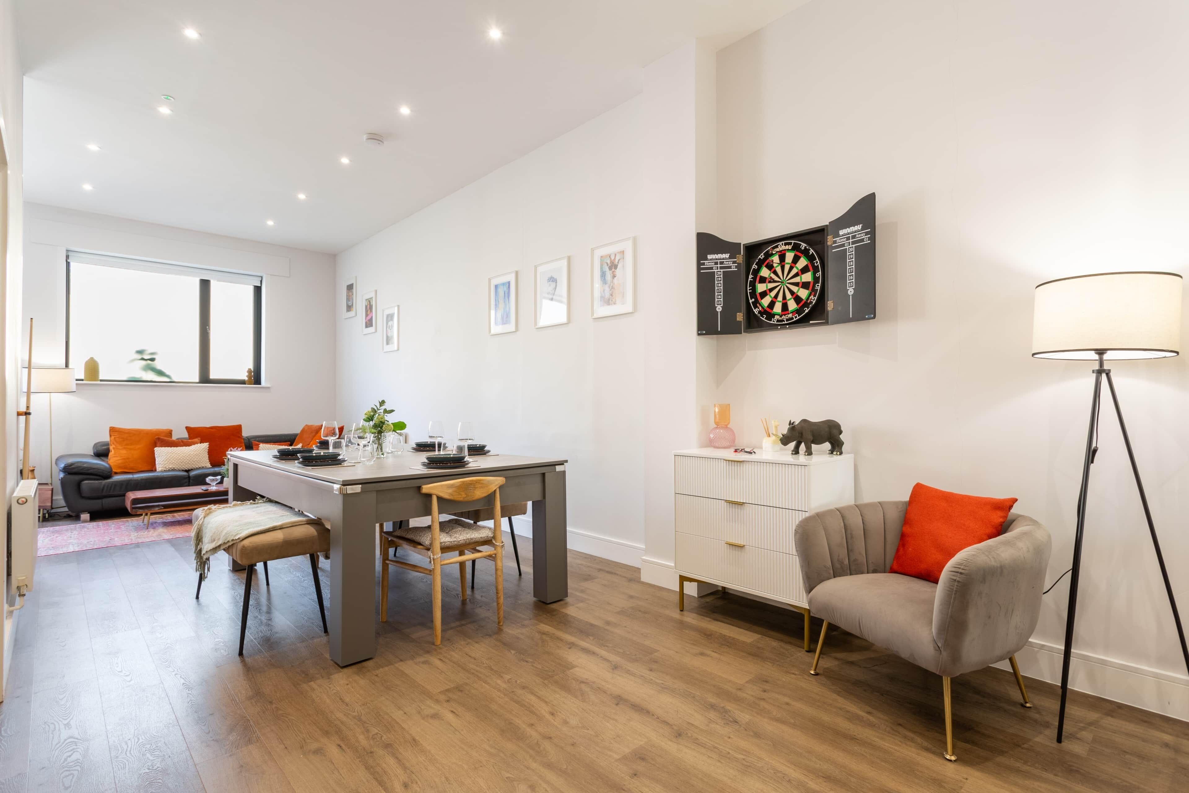 A modern living room with a dining table, a sofa, a lamp, and a dartboard on the wall.