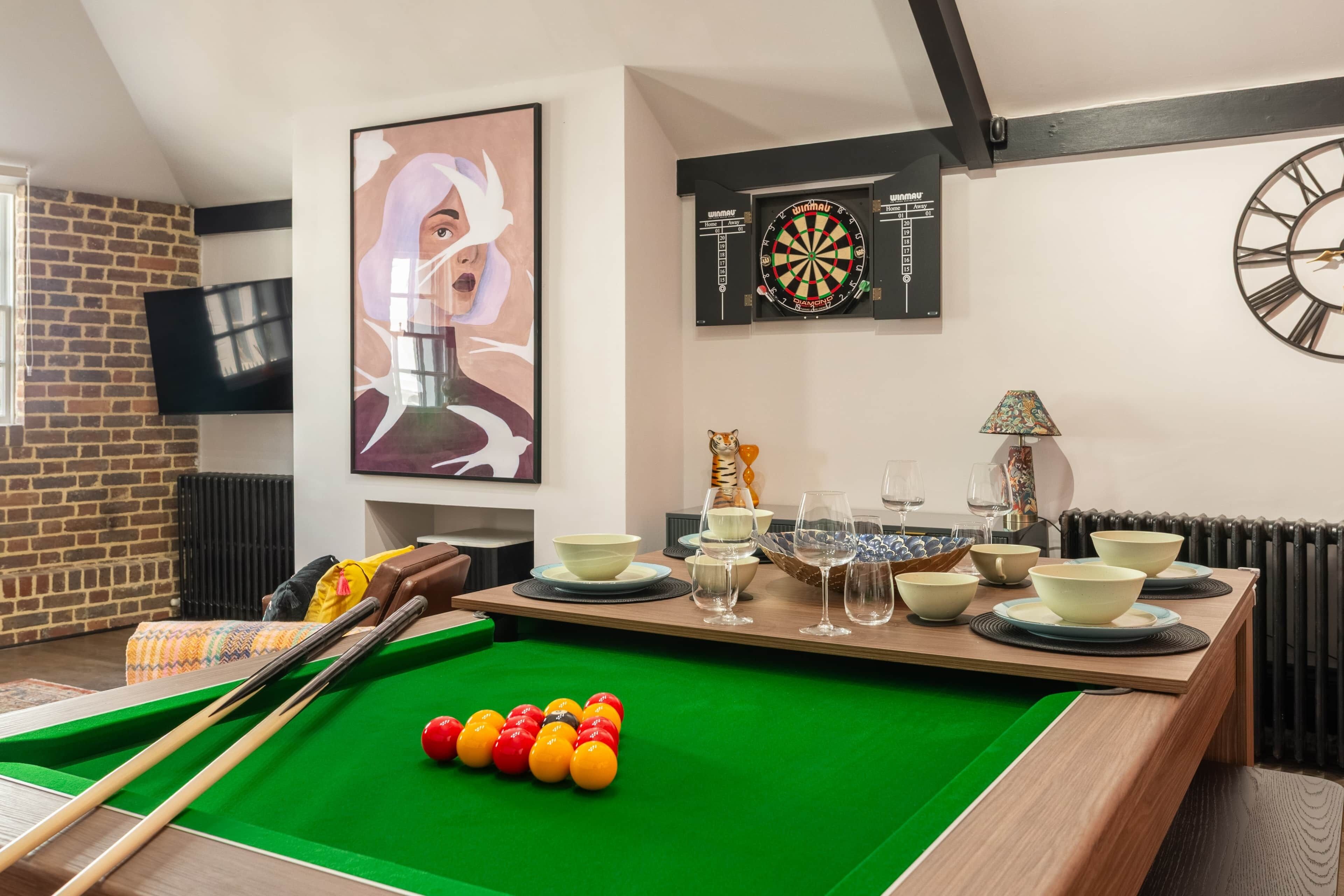 A billiards table with balls, a dartboard, a clock, and a framed artwork.