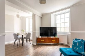 A modern living room with a dining area, a flat-screen TV on a wooden cabinet, and a blue sofa with dragonfly patterns. - Stylish 2-Bed 2- Bath Seafront Patio Apartment (Brighton and Hove)