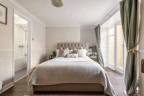 Un dormitorio con una cama grande, un cabecero, una mesita de noche, una ventana con cortinas y una puerta que da a un baño.