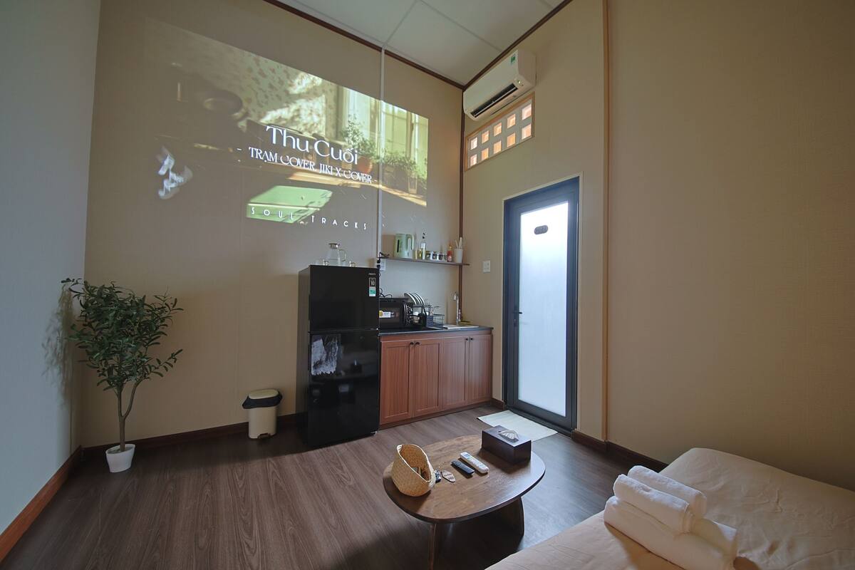 Hotelkamer met een bed, een tafeltje met een mand en afstandsbedieningen, een potplant, een kitchenette en een projectiescherm waarop 'Thuc Cuoi' staat.