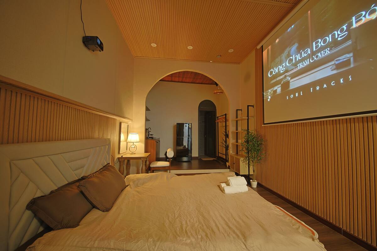 Een hotelkamer met een groot bed, een televisiescherm waarop een programma wordt getoond, een houten plafond en uitzicht op de gang.