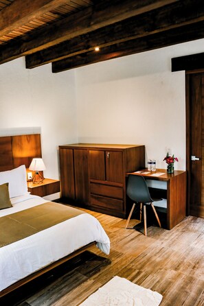Un dormitorio con cama, escritorio de madera, silla, mesita de noche, lámpara y una pequeña mesa con objetos decorativos.