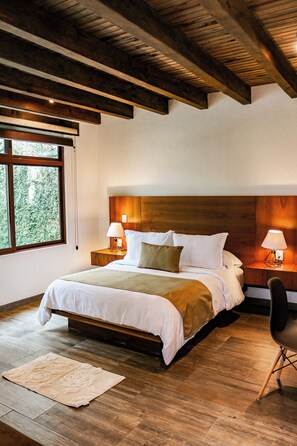 Un dormitorio con techo de madera, una cama grande con ropa de cama blanca, dos lámparas de noche, una silla y vistas a zonas verdes a través de una ventana.
