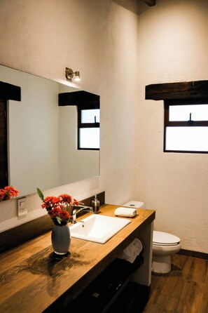 Un baño con tocador de madera, lavamanos blanco, espejo, inodoro y ventana.
