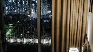 A hotel room with a bed, a nightstand with a lamp, and a view of a cityscape through a window with curtains. - Studio Suite Deluxe 35m2 with Balcony (Hà Nội)