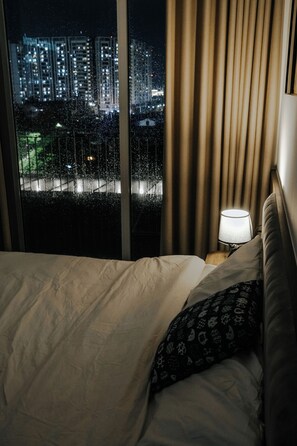 A hotel room with a bed, a nightstand with a lamp, and a view of a cityscape through a window with curtains. - Studio Suite Deluxe 35m2 with Balcony (Hà Nội)