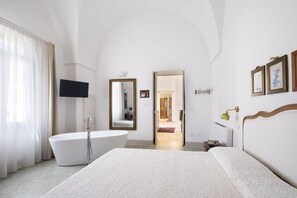 A modern bedroom with a large bed, a freestanding bathtub, a flat-screen TV, and a door leading to a bathroom.