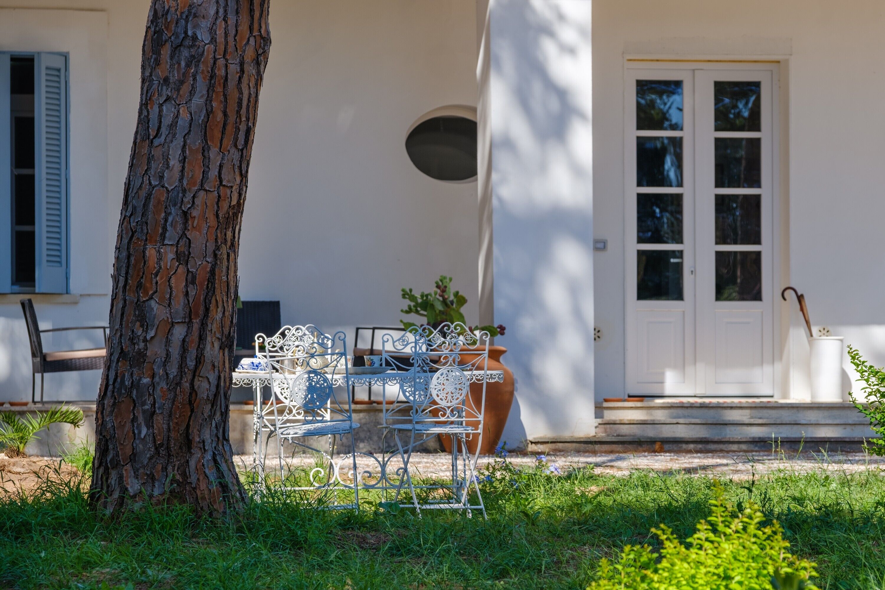Un edificio blanco con un árbol, una mesa de metal con sillas y una planta en maceta.